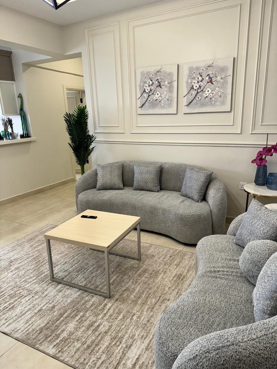 A light-filled seating area features a soft, curved gray sofa and a matching armchair, complemented by a simple wooden coffee table. Two floral artworks adorn the wall above, while a small potted plant adds a touch of greenery in the corner.