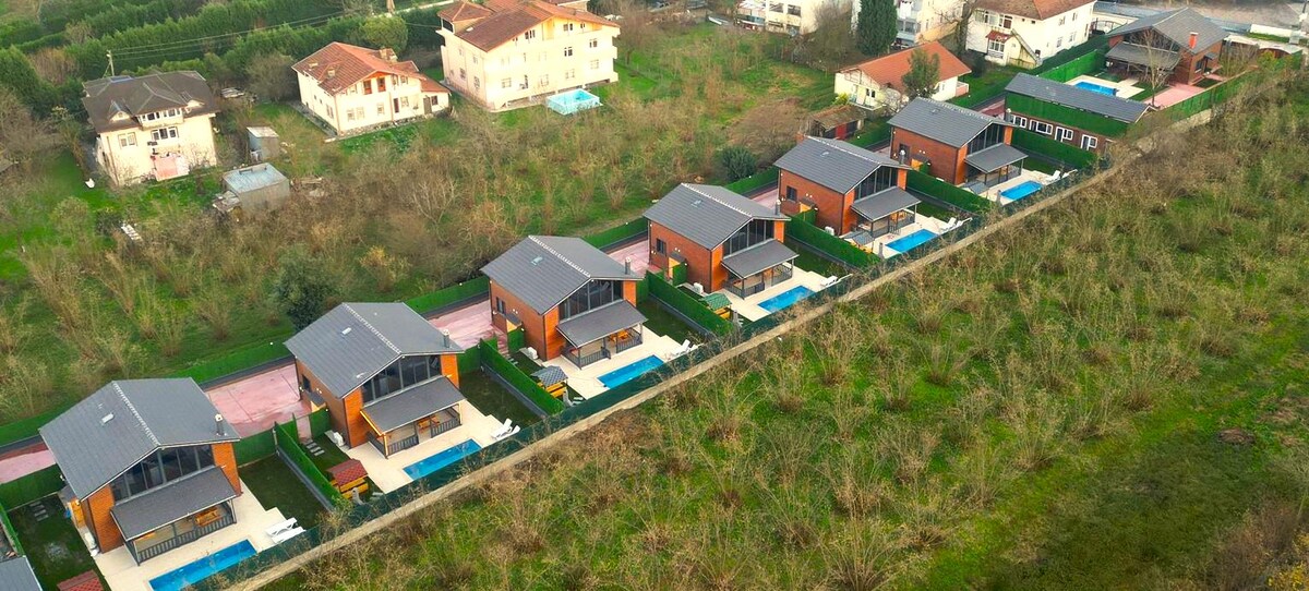 Aerial view of a resort featuring six identical houses, each with private pools and gardens. The landscape is surrounded by greenery, with pathways connecting the homes. Access to amenities is visible in the background, providing a sense of spaciousness and privacy.