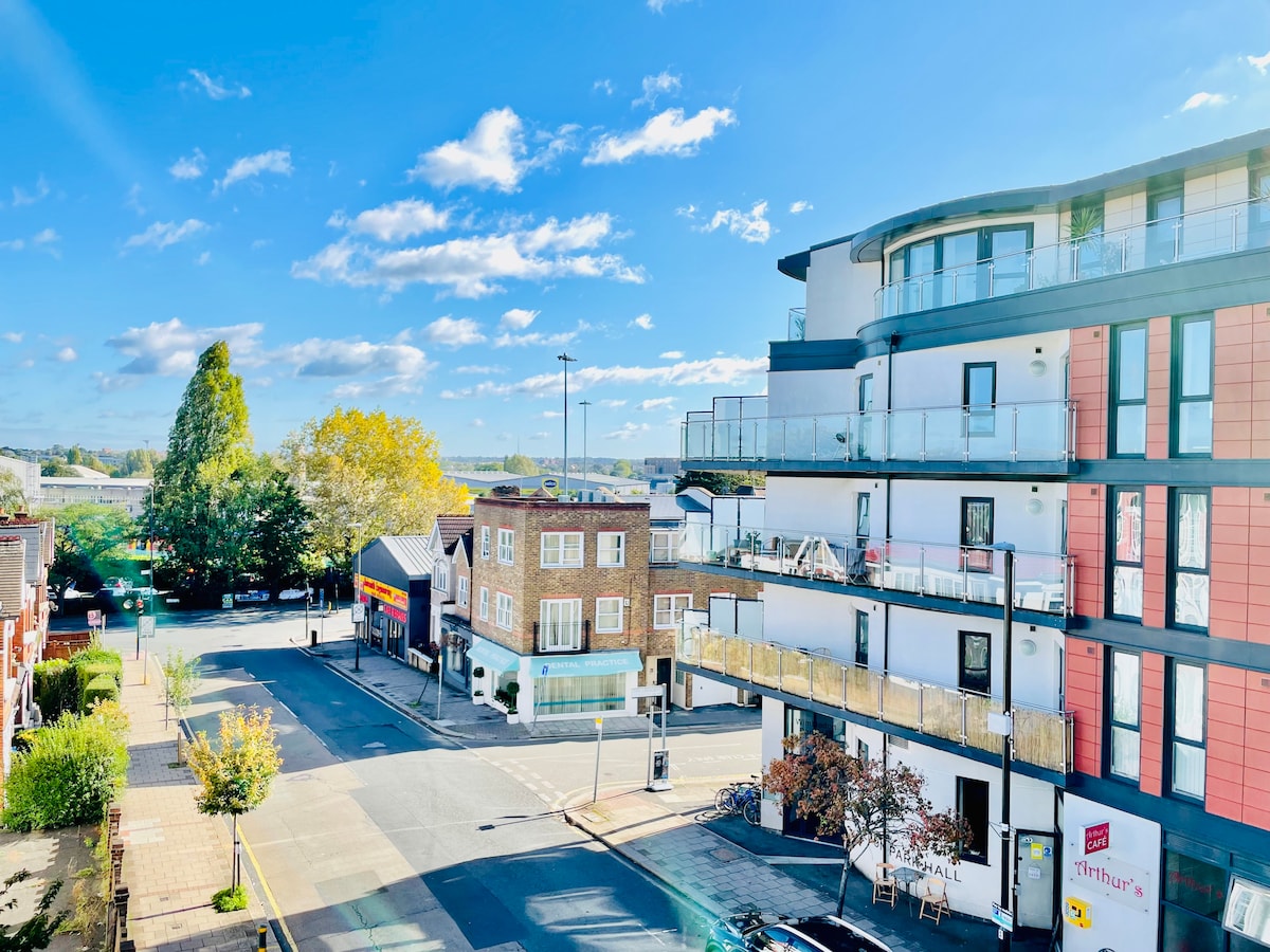 A view from the apartment showcases a spacious street lined with trees and a mix of brick and modern buildings. Bright blue skies and fluffy clouds highlight the tranquil atmosphere of the neighborhood, while the vibrant colors of the structures create a welcoming urban scene.