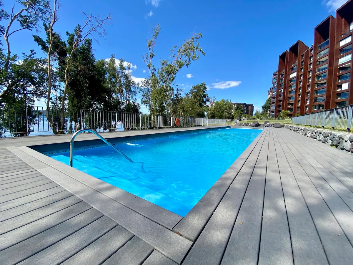 A clear blue swimming pool is framed by wooden decking, offering an inviting space for relaxation. Lush greenery surrounds the area, with trees providing shade. In the background, modern buildings can be seen, highlighting the scenic view of the nearby lake.