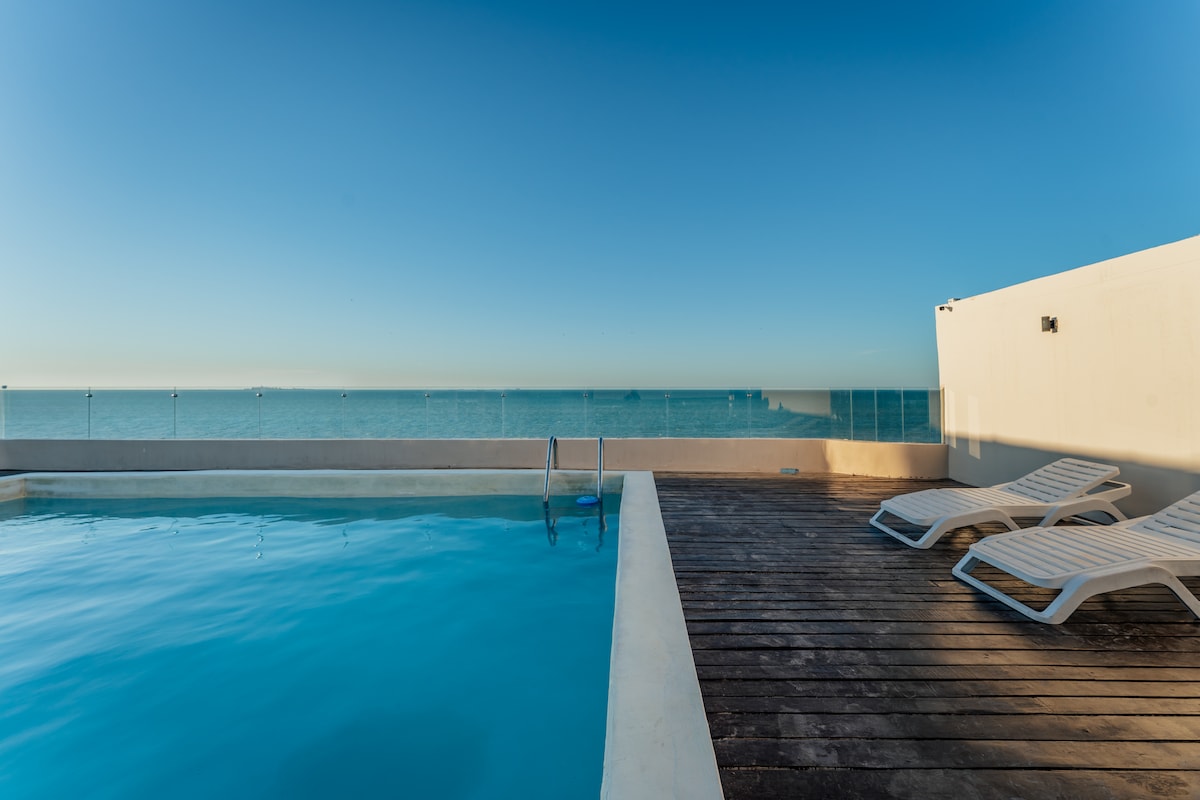 A rooftop area is shown, featuring a sparkling swimming pool with clear blue water. Comfortable lounge chairs are arranged around the pool on a wooden deck, providing an unobstructed view of the sea and sky, creating a serene environment for relaxation.