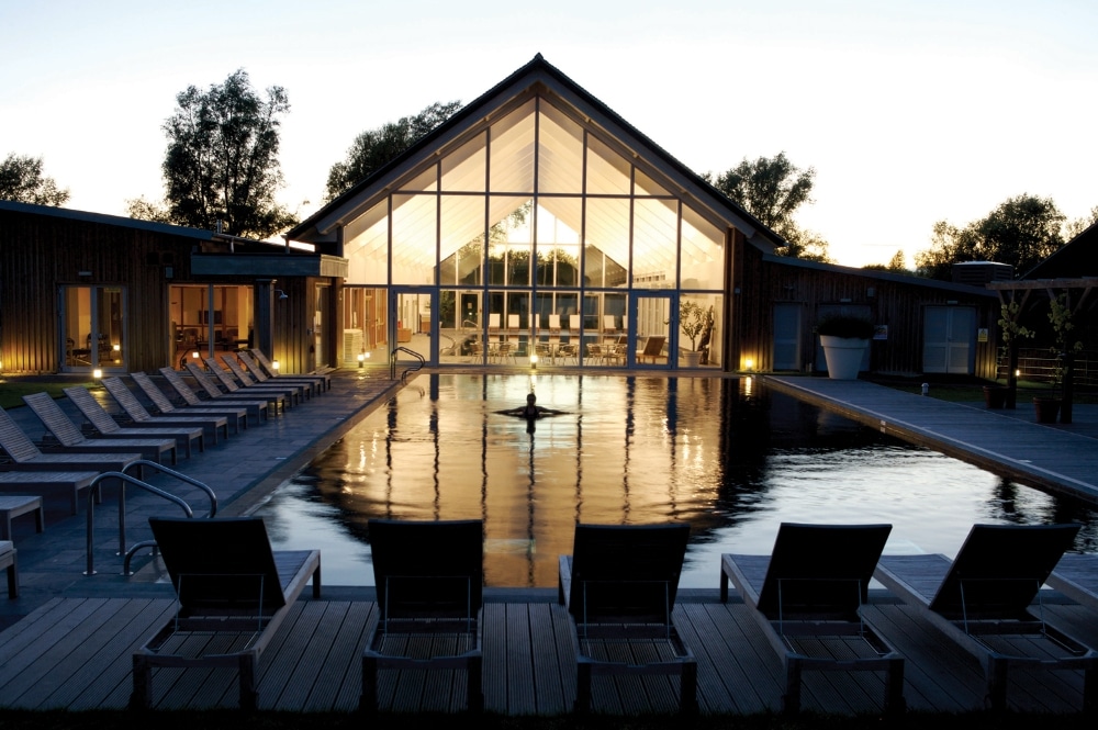 A modern, spacious spa facility is depicted, featuring large glass paneling that reflects evening light. An outdoor pool is illuminated, bordered by lounge chairs arranged neatly along the deck, creating an inviting area for relaxation.