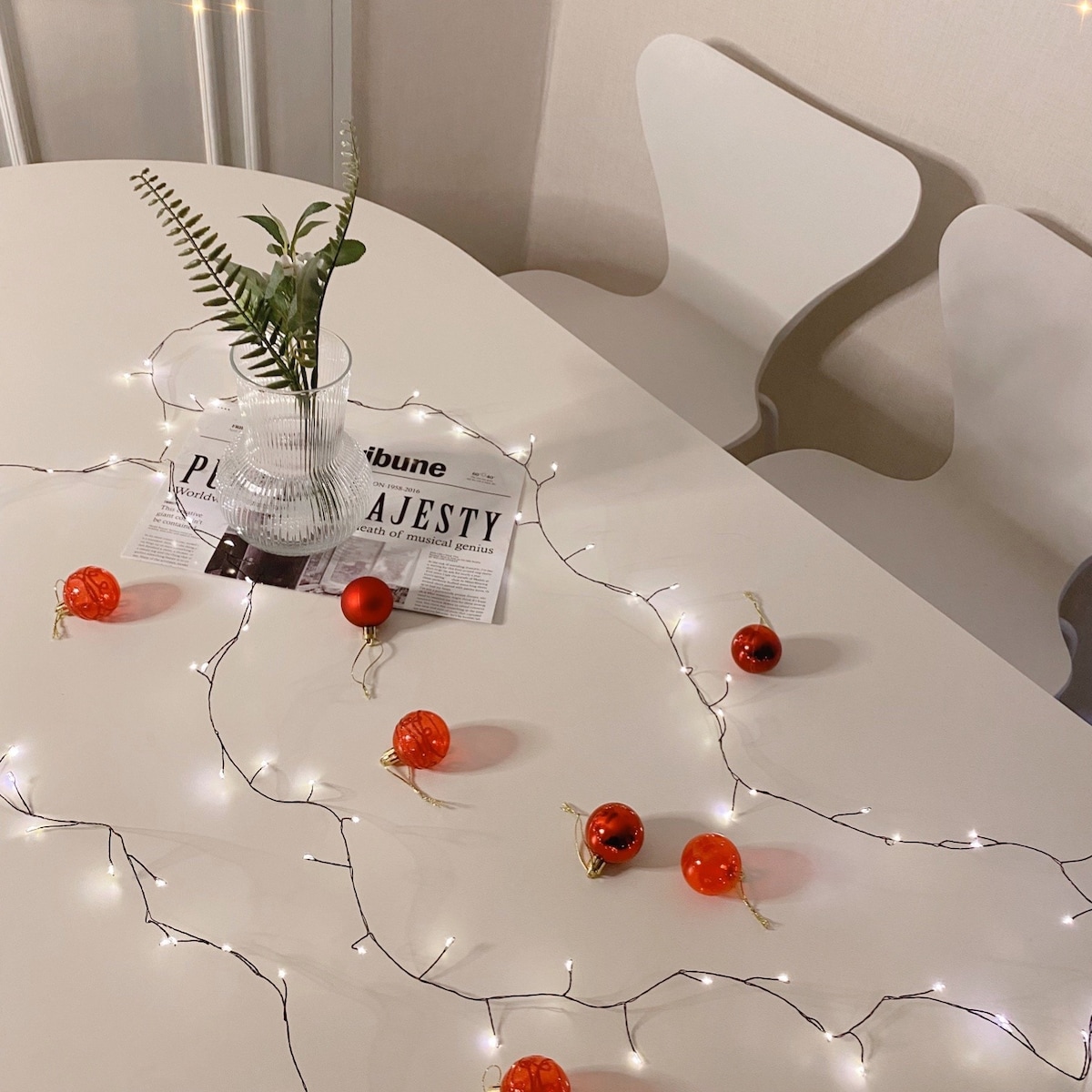 A white table is adorned with delicate fairy lights and small orange decorations. A glass vase containing a green plant is placed at the center, complemented by a magazine. Two modern white chairs are partially visible in the background.