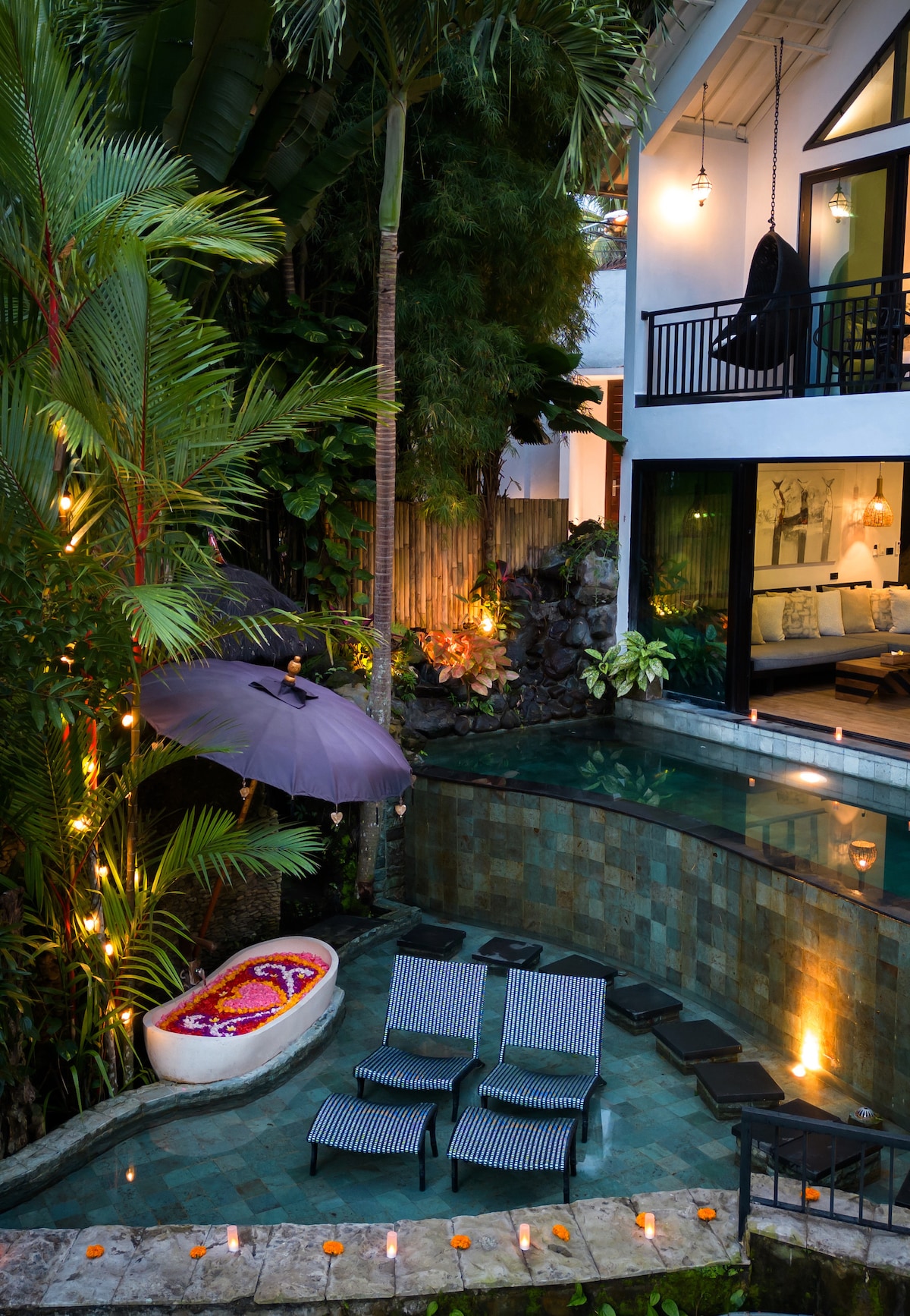 An outdoor space showcases a private greenstone pool surrounded by lush tropical foliage. Two lounge chairs are positioned near the water, accompanied by a decorative umbrella. Soft lighting enhances the serene atmosphere, and a wooden alcove offers a cozy seating area visible through expansive glass doors.
