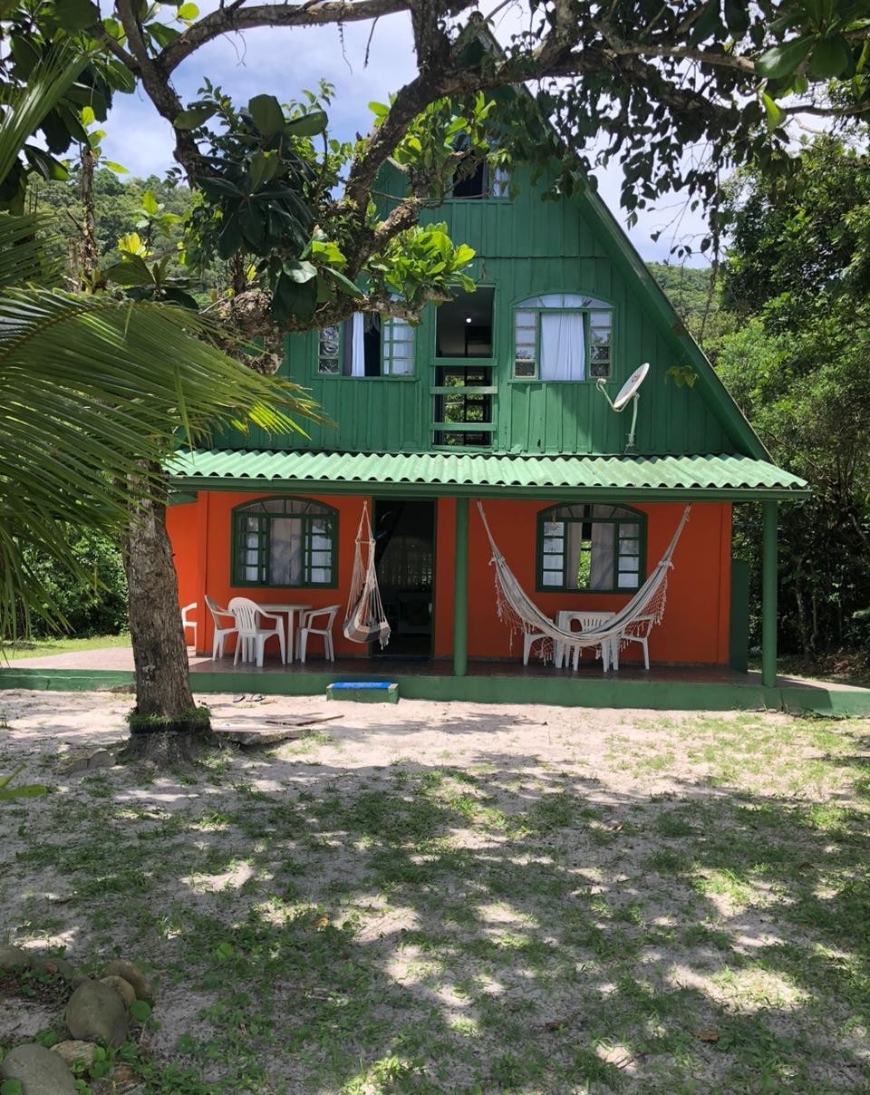 A two-story chalet features a vibrant orange and green exterior. Several large windows and a sloped roof enhance its charm. In the foreground, white chairs and hammocks are arranged under the shade of nearby trees, providing a comfortable outdoor space.