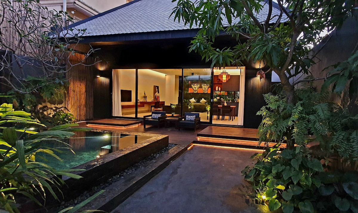 The exterior of a tropical villa is framed by lush greenery, featuring a private salt-water pool and wooden deck. Expansive glass doors provide views into the open-plan living area, illuminated by warm lighting, enhancing the inviting atmosphere of the courtyard.