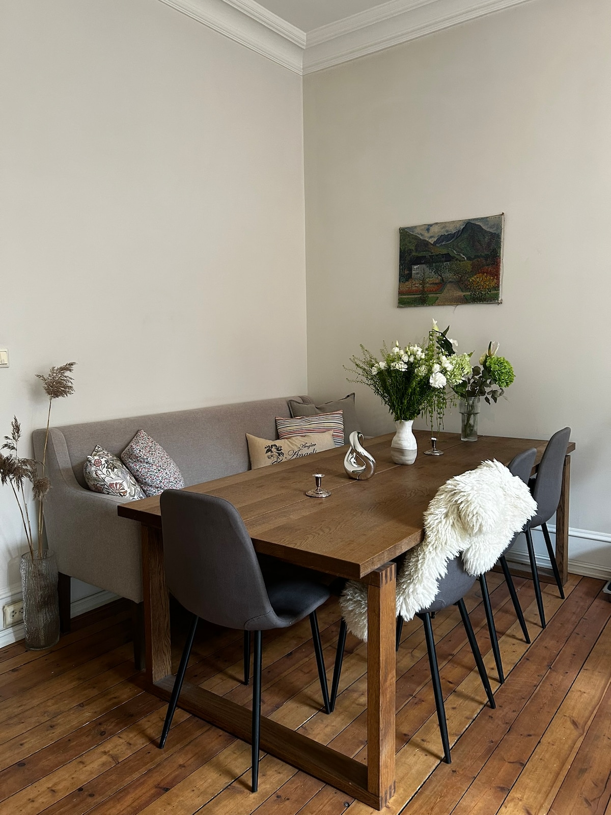 A dining area features a wooden table accompanied by four chairs and a cozy bench. Decorative pillows adorn the bench, while a vase of fresh flowers sits at the center. A piece of artwork is displayed on the wall, adding character to the space.
