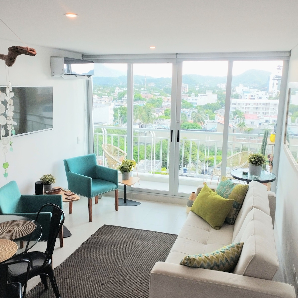 A bright living area features large glass windows offering views of the surrounding area. A comfortable white sofa is paired with teal armchairs and a round dining table. Decorative plants are placed strategically, enhancing the space's openness and connection to the outdoors.