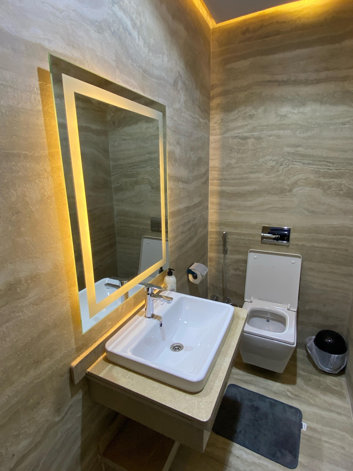 A well-appointed bathroom features a modern sink below a softly illuminated mirror. The walls display a natural stone pattern, complemented by a toilet and a waste bin. A bath mat is placed on the floor, enhancing comfort and functionality.