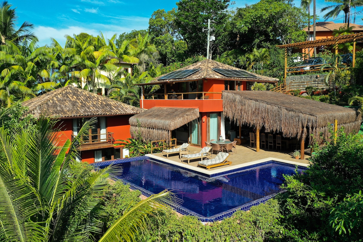 A spacious villa is nestled among lush greenery, displaying a large outdoor pool surrounded by palm trees. The thatched-roof areas provide shaded seating. Brightly colored walls complement the natural surroundings, offering a harmonious blend of architecture and landscape.