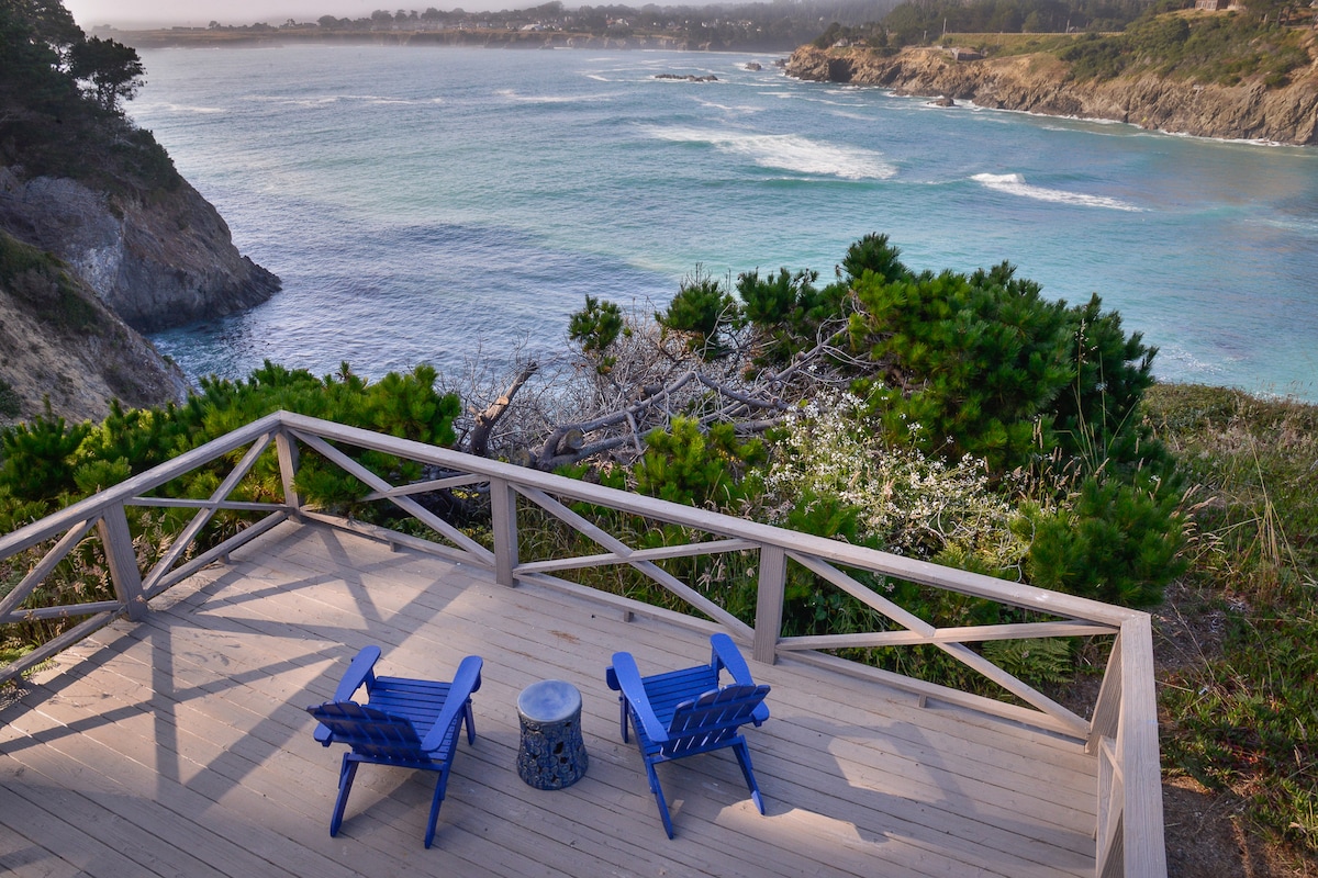 A private deck offers two blue chairs positioned to overlook the ocean. The natural landscape includes green shrubs and rocky terrain leading to the water's edge. Gentle waves can be seen lapping at the shore, with inland views of the distant village and coastal areas.