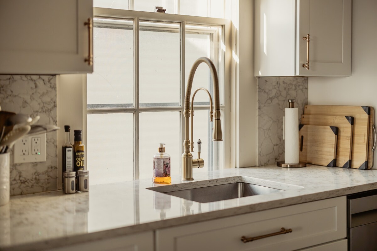 A modern kitchen features light-colored cabinets and a marble countertop. A sleek gold faucet stands above a deep sink, with a window providing natural light. Wooden cutting boards are arranged on the counter, alongside bottles of oil and vinegar, enhancing the functional space.