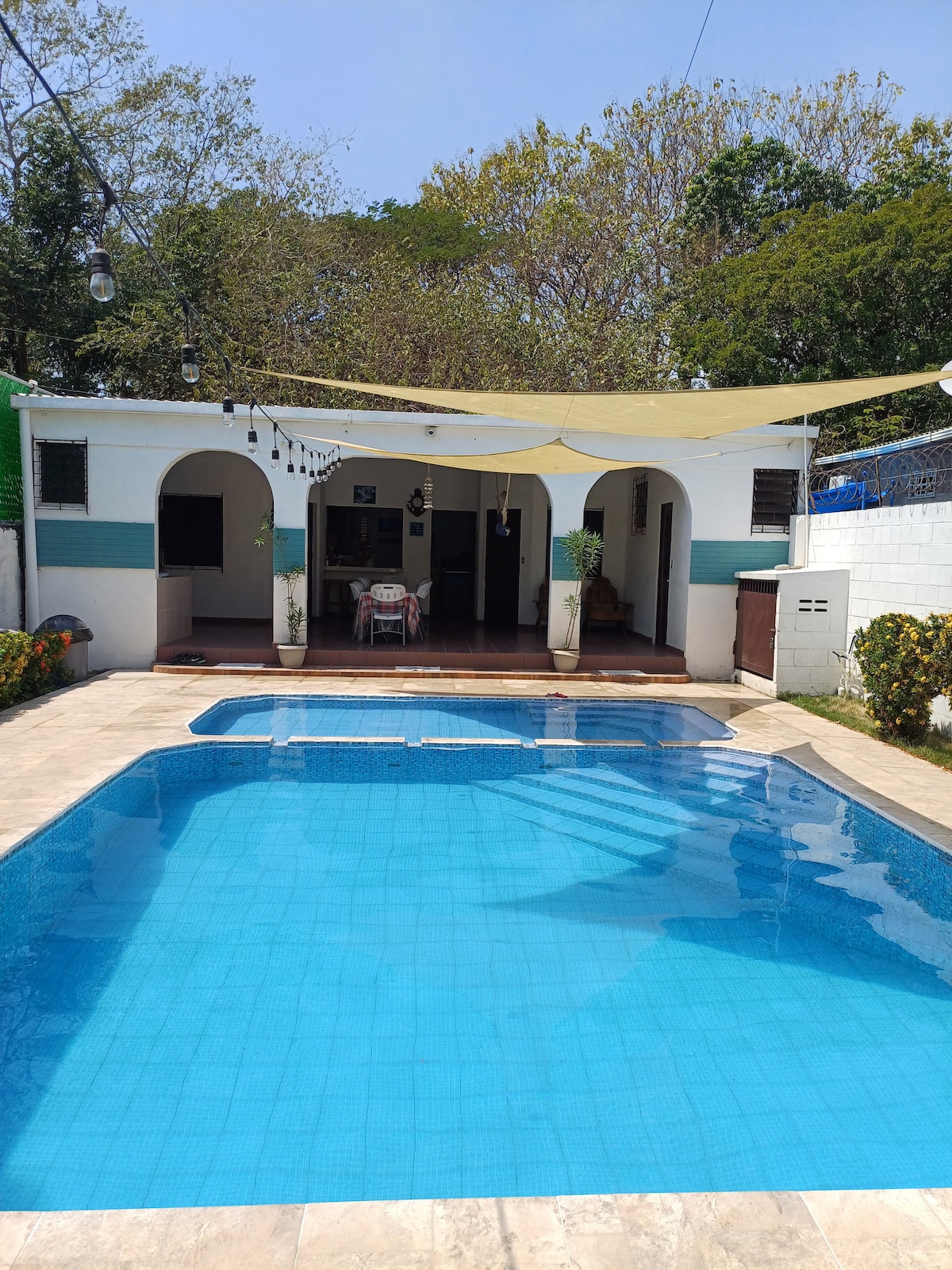 A refreshing pool is framed by a tiled patio, with two distinct areas for swimming. A shaded seating area is visible in the background, surrounded by green plants. The house's exterior features arched entrances, providing a relaxed atmosphere and a connection to the outdoor space.