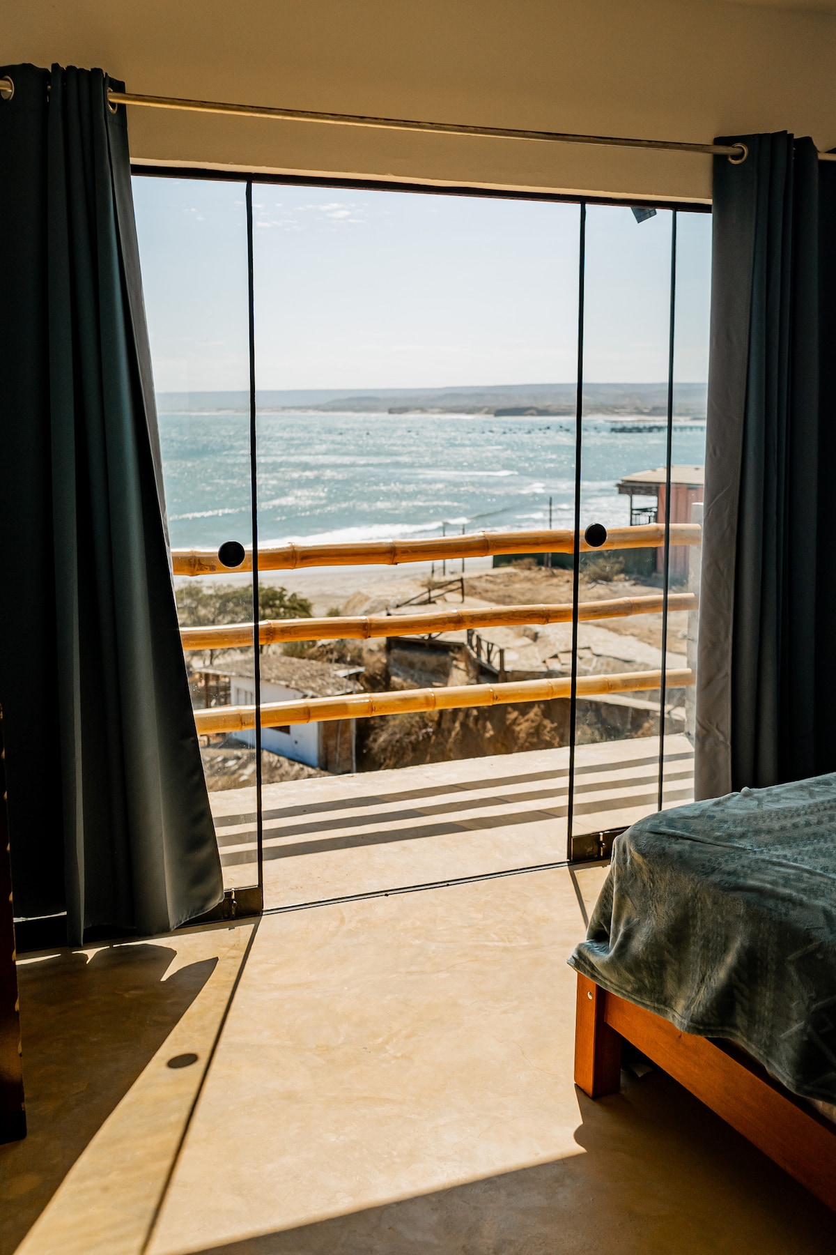 A sliding glass door opens to a balcony, revealing sweeping ocean views. Soft natural light fills the space, highlighting the wooden bed. Dark curtains frame the door, complementing the beachside setting.