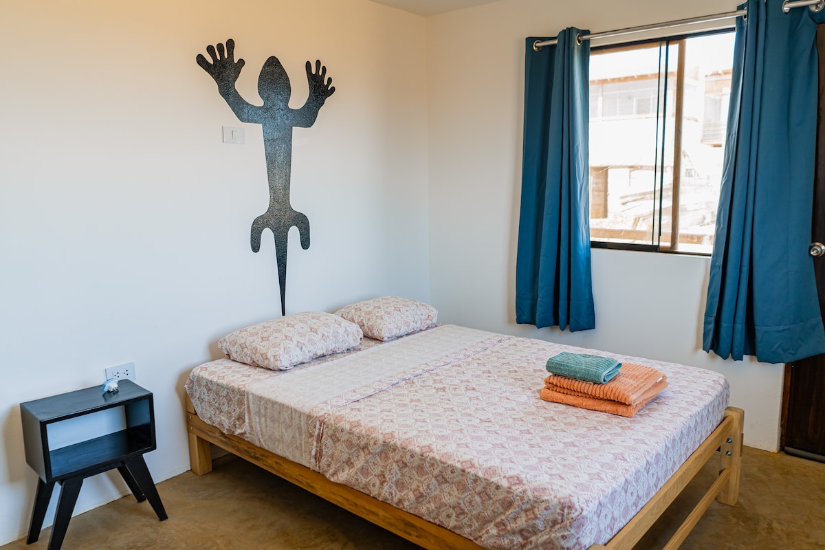 A comfortable sleeping area features a full-size bed adorned with patterned bedding and neatly arranged towels. A wooden nightstand sits next to the bed, while a window dressed with blue curtains allows natural light to fill the space. A decorative wall piece is displayed above the bed.