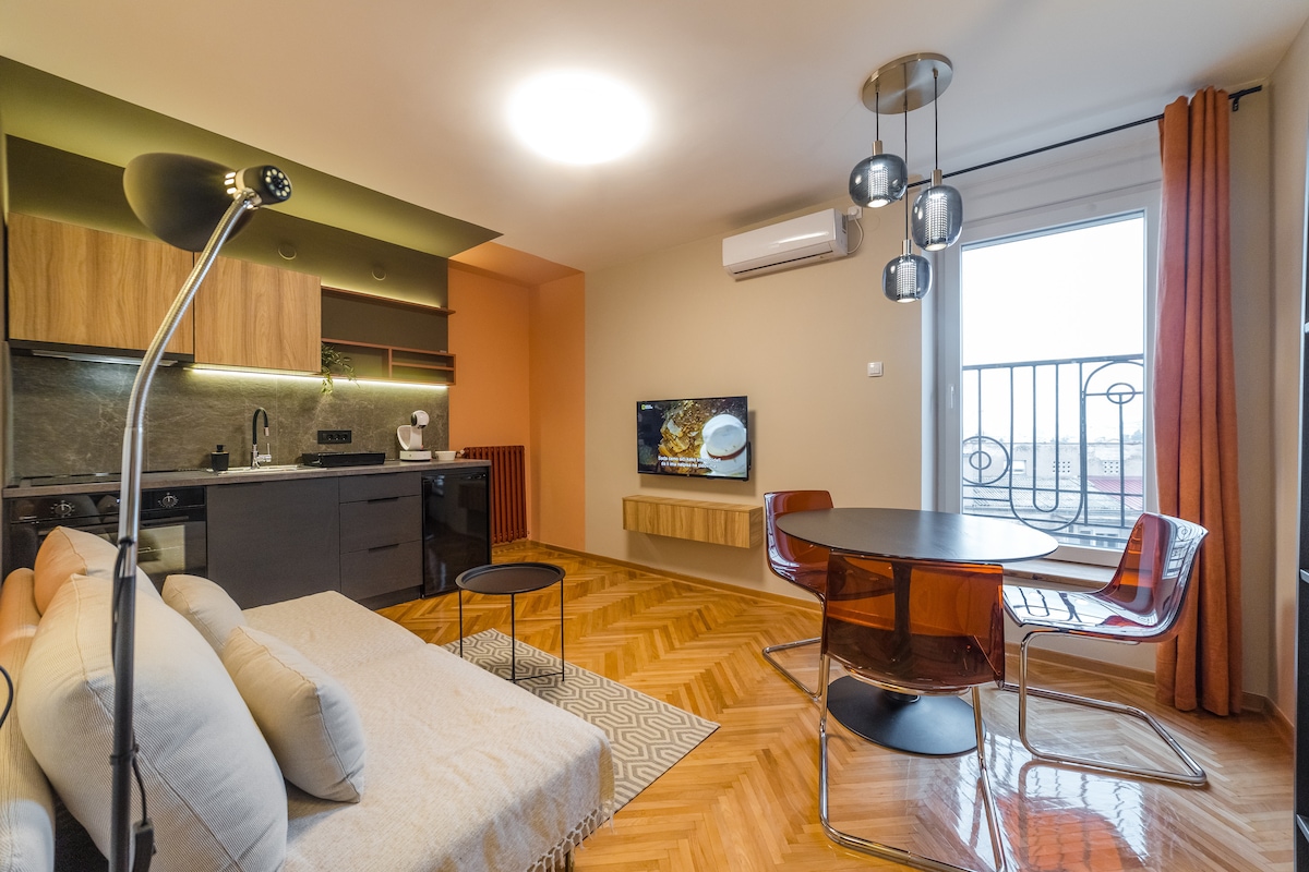 A modern living space features a mix of warm wood cabinetry and sleek grey countertops. A circular dining table with chairs is situated near the large window, which provides natural light. A flat-screen TV is mounted on the wall, and a soft sofa adds comfort to the room.
