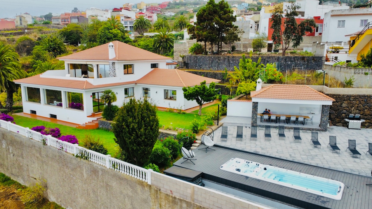 The villa showcases a modern architectural design set within a spacious garden, featuring vibrant shrubs and palm trees. A large outdoor area includes a heated jacuzzi and barbecue, surrounded by seating. The serene landscape offers glimpses of distant residential buildings and greenery.