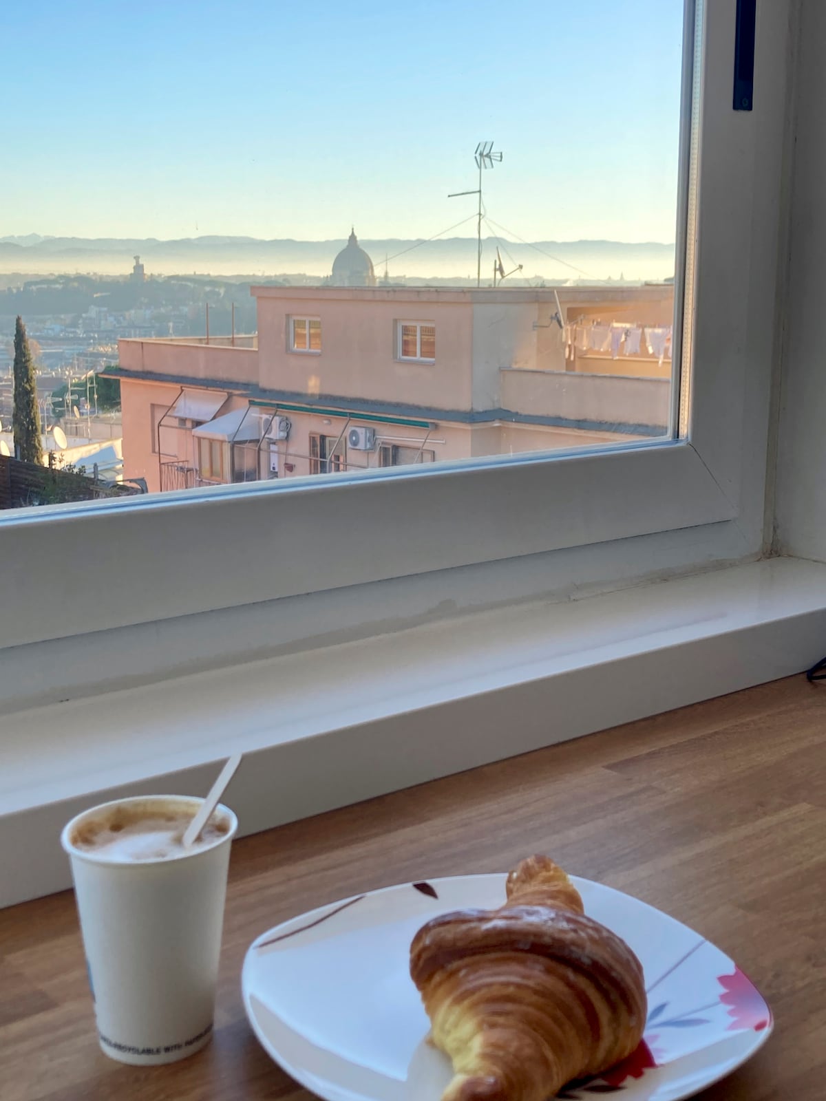 A bright window offers a panoramic view of distant hills and the skyline of Rome, featuring the dome of St. Peter's Basilica. A cup of coffee and a croissant are positioned on a wooden surface, suggesting a serene morning setting.