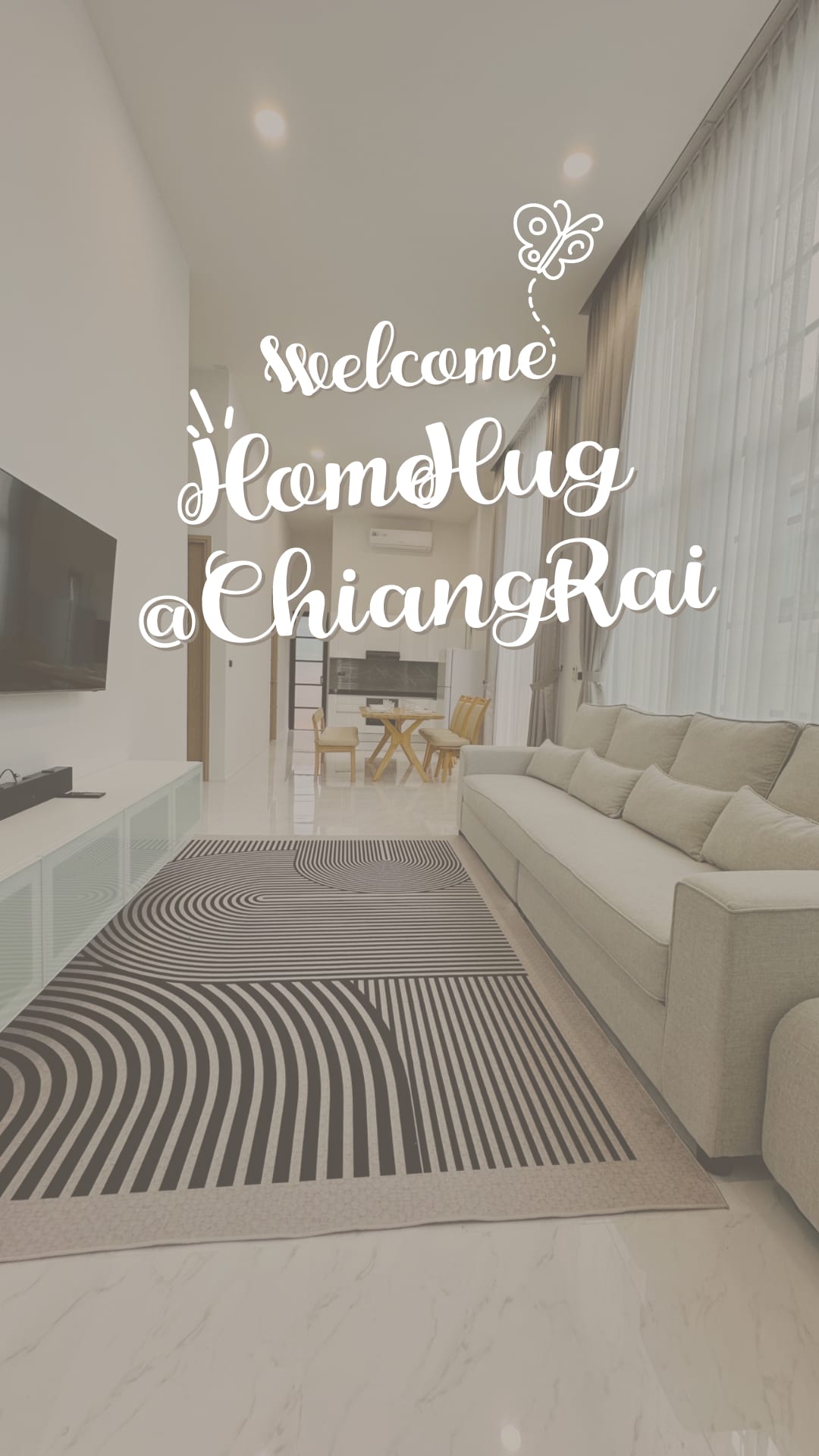 A modern living area is presented with a spacious sofa, complementing neutral-toned walls. A large flat-screen TV is mounted on the wall opposite a patterned rug. Natural light filters through tall windows, creating an open and airy feel in the space.