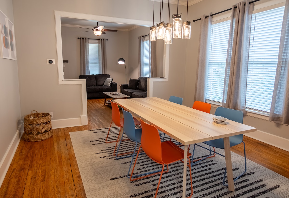A dining area is presented with a light wood table surrounded by modern orange and blue chairs. Soft light is provided by hanging fixtures above. Large windows with light-filtering curtains allow natural light to fill the space, complementing the neutral walls and warm wood floor.