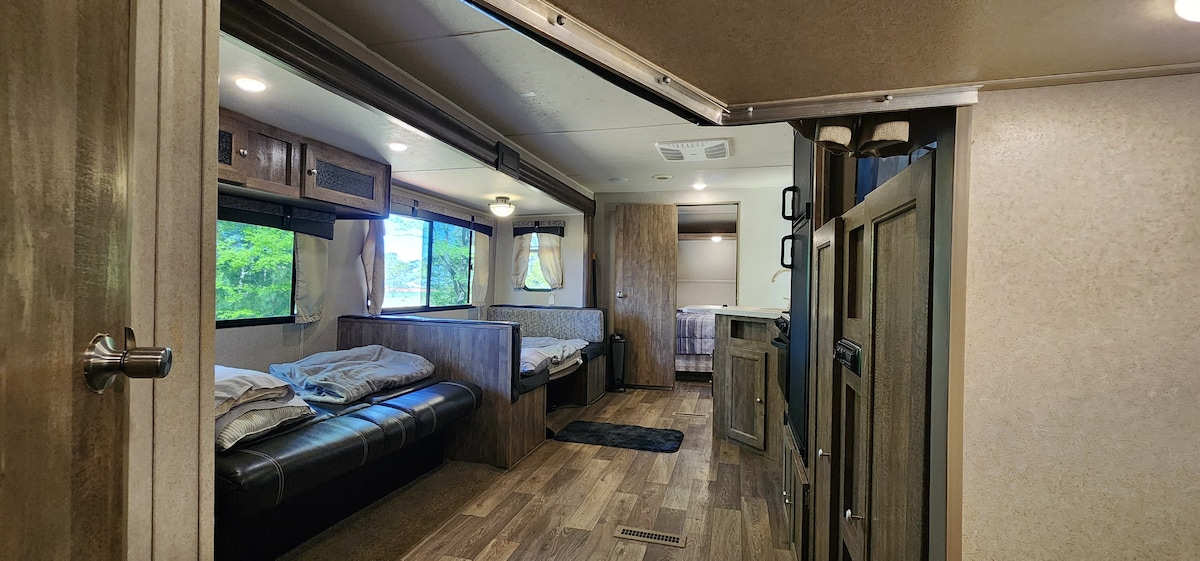 The interior of a camper features a cozy seating area with dark leather couches and natural wood accents. Large windows allow natural light to fill the space, highlighting a comfortable dining nook with bench seating. A soft area rug sits on the wooden floor, adding warmth to the environment.