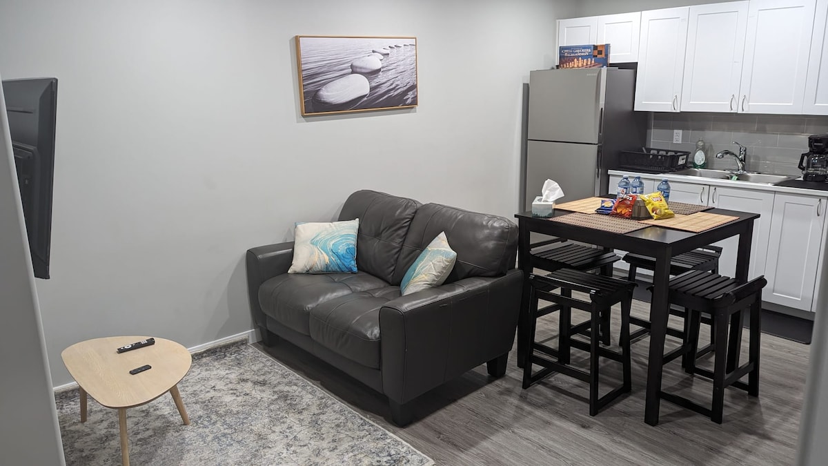 A living area displays a dark leather sofa adorned with light blue and cream cushions. A minimalist coffee table sits in front, while a compact dining set with black stools occupies a corner. A modern kitchen is visible in the background, featuring a refrigerator and additional appliances.