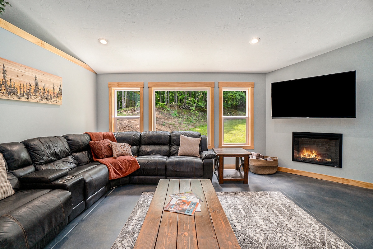 The living area features a spacious black leather sectional sofa, accompanied by decorative pillows and a cozy throw. Natural light fills the room through large windows, and a modern television is wall-mounted above a gas fireplace. A rustic-style coffee table sits on a textured area rug.
