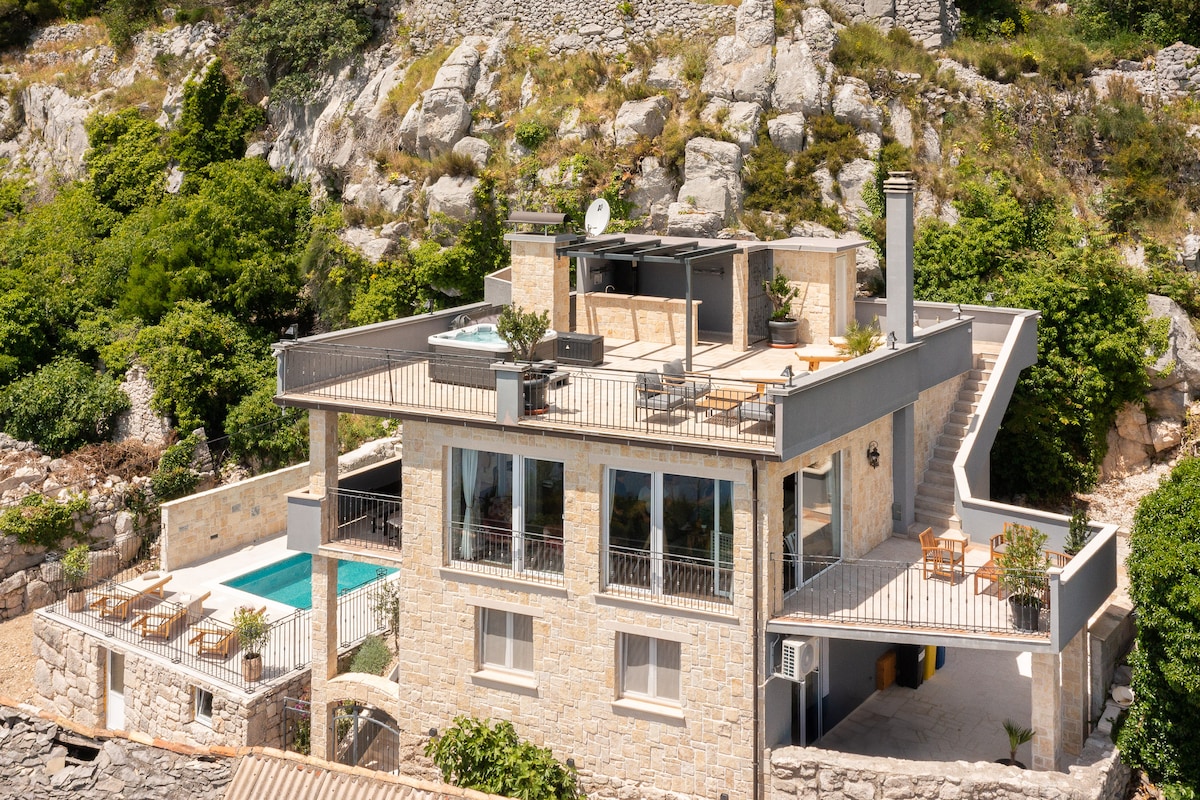 A modern stone house is nestled among greenery, featuring multiple terraces with outdoor seating. A private heated swimming pool is visible, along with a jacuzzi on the upper level. The structure's design highlights extensive windows, maximizing natural light and showcasing scenic views.