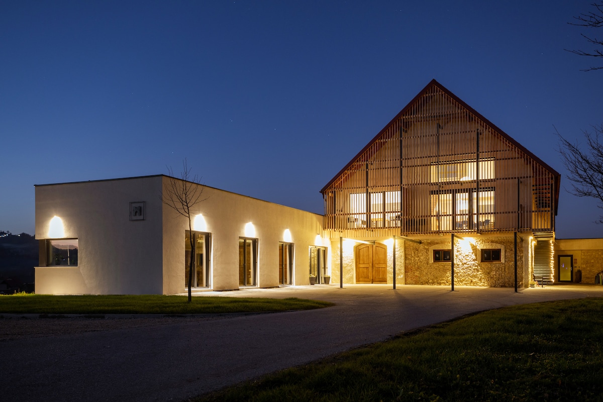 The exterior of the property is illuminated at night, showcasing a spacious, two-story building with a wooden slatted balcony. Warm lights accentuate the stone and white stucco, creating a welcoming atmosphere. A manicured lawn borders the pathway leading to the main entrance.