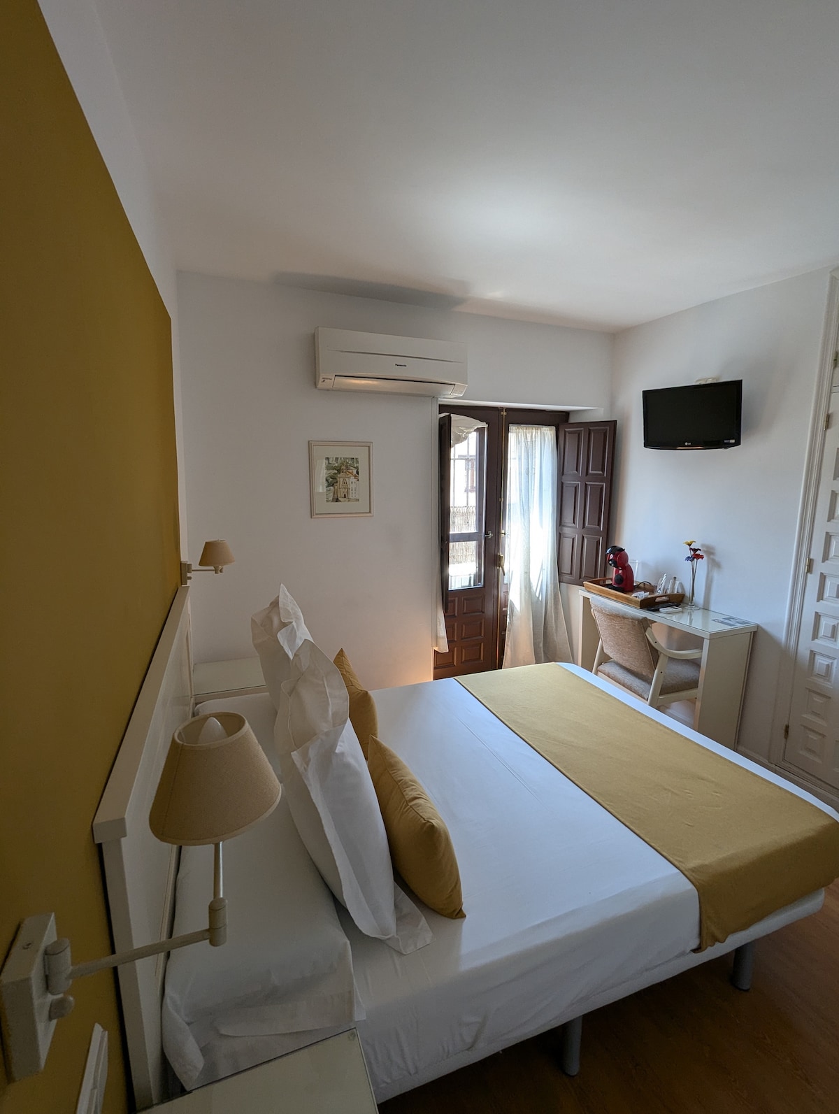A bright room features a spacious bed with a light-colored blanket and decorative pillows. A small desk and chair are positioned near a window, alongside a wall-mounted television. The door opens to a small balcony, allowing natural light to fill the space.