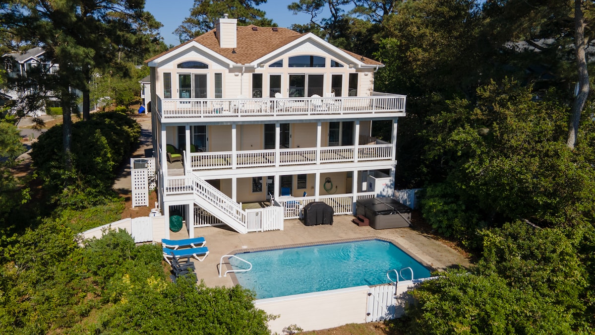 A spacious sound-front home is showcased, featuring multiple levels with expansive decks. A private pool is visible in the foreground, surrounded by lounge chairs. Lush greenery frames the property, and the backdrop includes trees and neighboring homes.