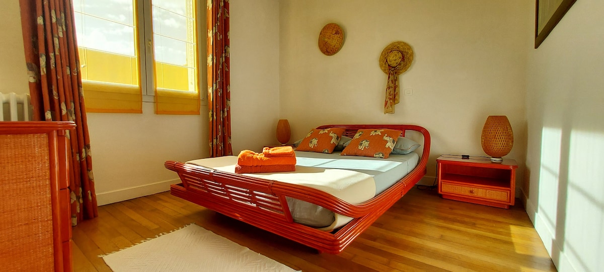 A brightly colored room features a spacious bed with light blue bedding adorned with floral patterns. Decorative orange towels are neatly arranged on the bed. Two woven wall hangings add texture, and natural light filters through large windows framed by patterned curtains, illuminating the wooden flooring.