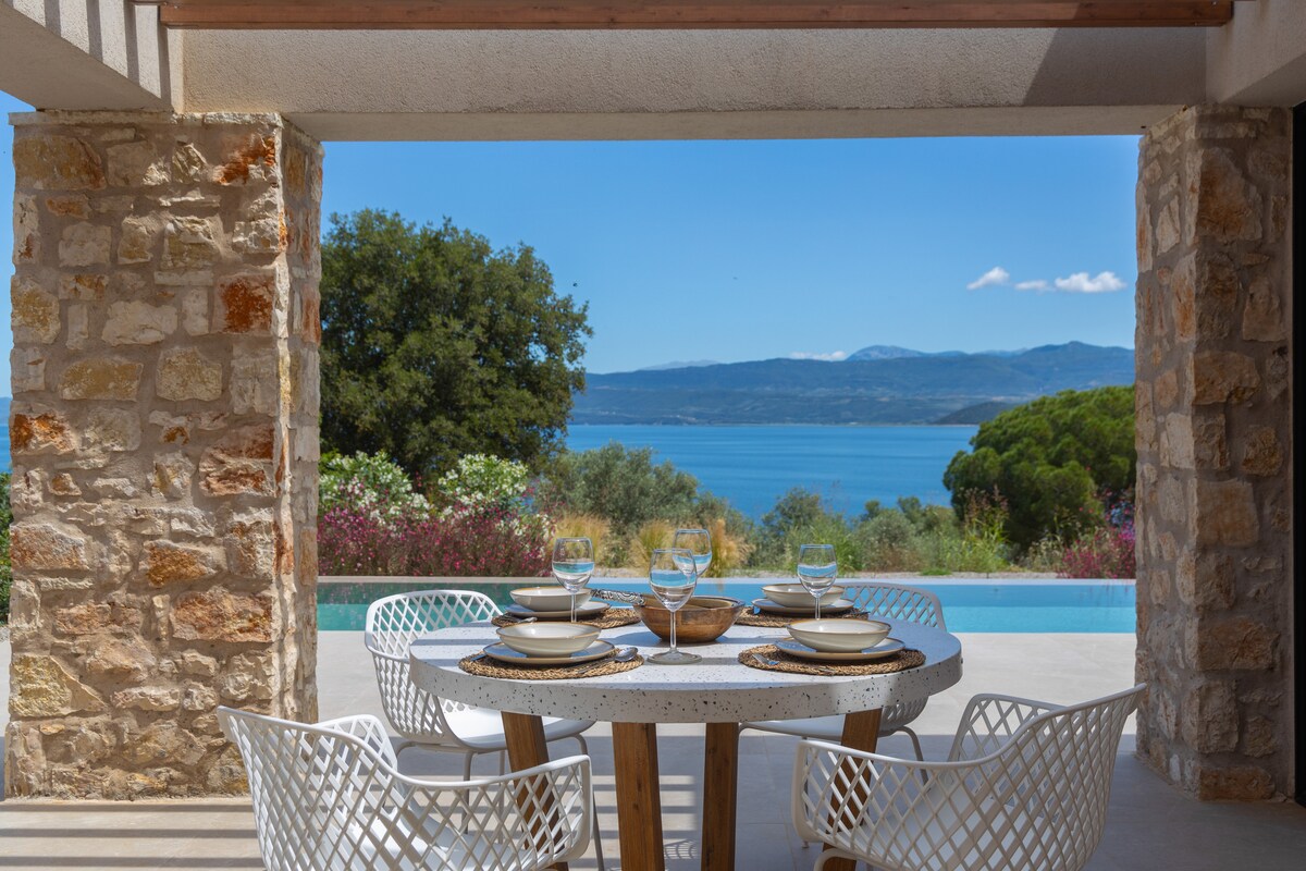 An outdoor dining area features a round table set with ceramic plates and utensils, surrounded by four white woven chairs. The scenic view of the Ambracian Gulf is visible through stone pillars, showcasing the tranquil waters and distant mountains under a clear blue sky.