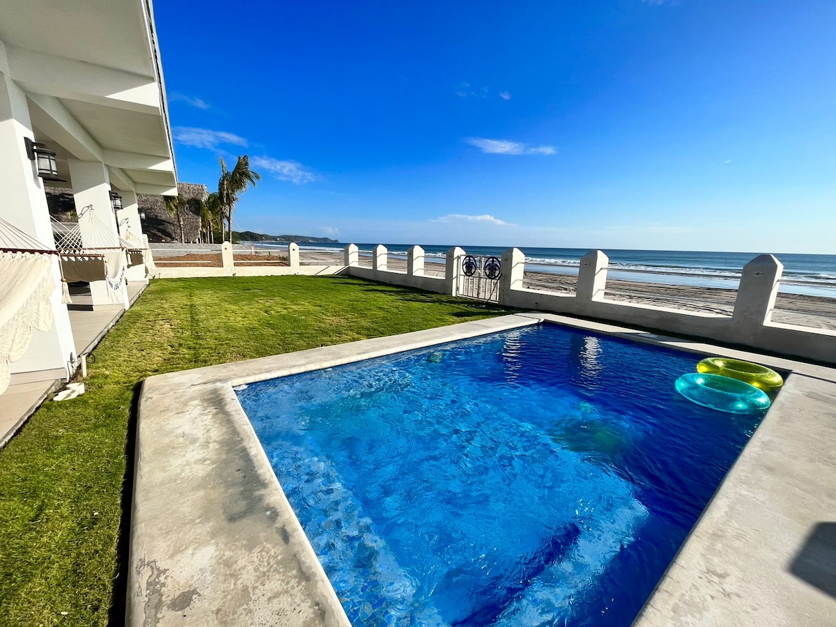 A private pool is surrounded by lush green grass, inviting relaxation on warm days. Two colorful floats rest on the clear water, while the ocean and sandy beach are visible in the background under a bright blue sky.