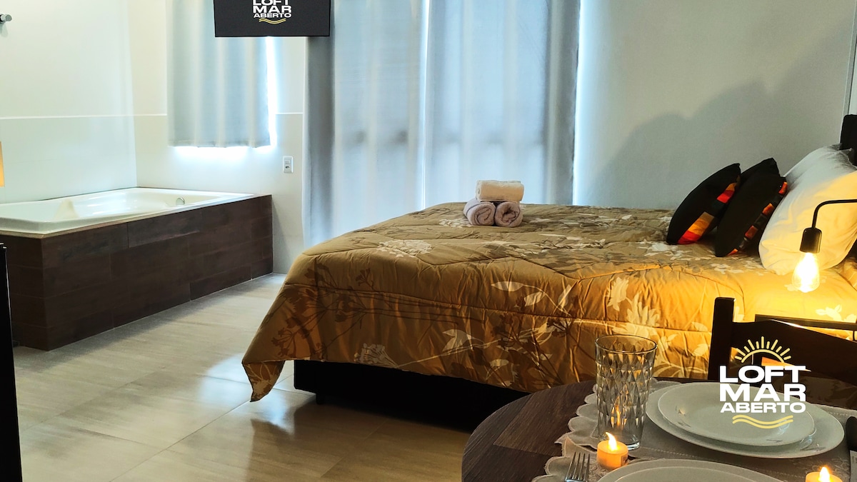 A cozy sleeping area is presented with a queen-sized bed featuring floral bedding and complimentary pillows. A large window is covered by sheer curtains, allowing soft light to enter. A stylish dining setup is visible beside the bed, along with a relaxing bathtub in the corner.