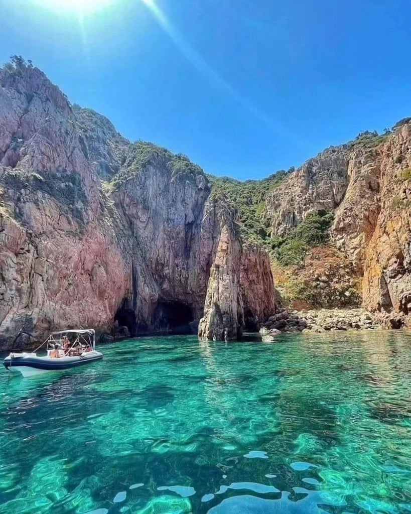A tranquil cove reveals steep rocky cliffs adorned with greenery. Crystal-clear turquoise waters gently lap at the shoreline, where a small boat is anchored near the base of the cliffs. Sunlight reflects off the water, creating a serene atmosphere.