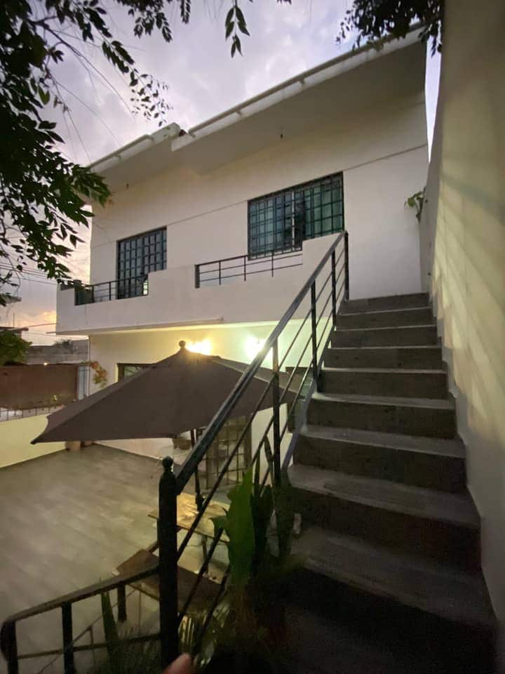 A two-story building is shown with a staircase leading to the upper level. Large windows are positioned on the façade, allowing for natural light. A patio area is visible with a shaded umbrella, surrounded by greenery, providing a serene outdoor space.