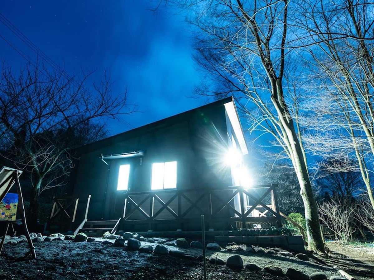 A modern guesthouse is illuminated against the night sky, featuring large windows and a wooden porch. Surrounding trees are softly lit, creating a serene outdoor ambiance. The pathway is lined with smooth stones, guiding the way to the entrance.