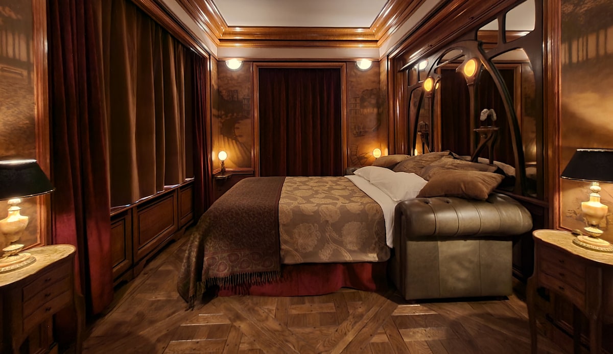 A cozy bedroom features intricate wood paneling and hand-carved Art Nouveau details. A spacious bed is dressed in patterned linens, complemented by soft lighting from bedside lamps. Rich velvet drapes frame the window, adding to the room's elegant, period-inspired atmosphere.