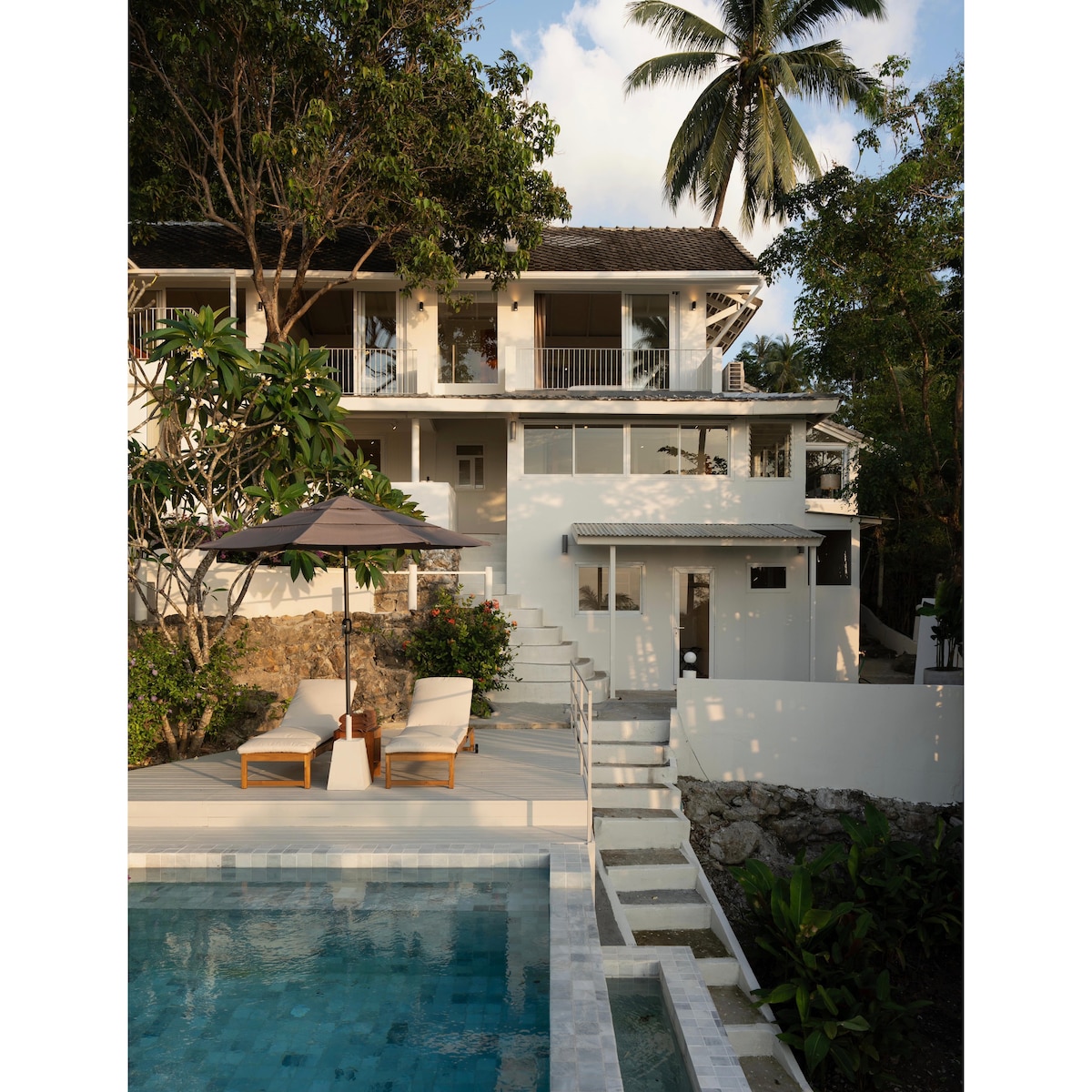 The image depicts a modern villa nestled among lush greenery, showcasing a spacious infinity pool in the foreground. Sun loungers with a small umbrella are positioned nearby, leading to a series of steps that ascend to a well-lit upper level of the villa.