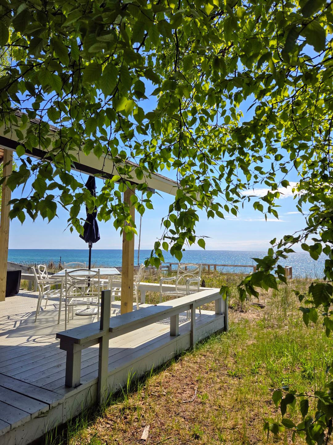 A covered deck is framed by lush green leaves, offering a view of Lake Michigan. White patio furniture is arranged for al fresco dining, while a large umbrella provides shade. The calm water sparkles under the bright blue sky, creating a serene atmosphere.