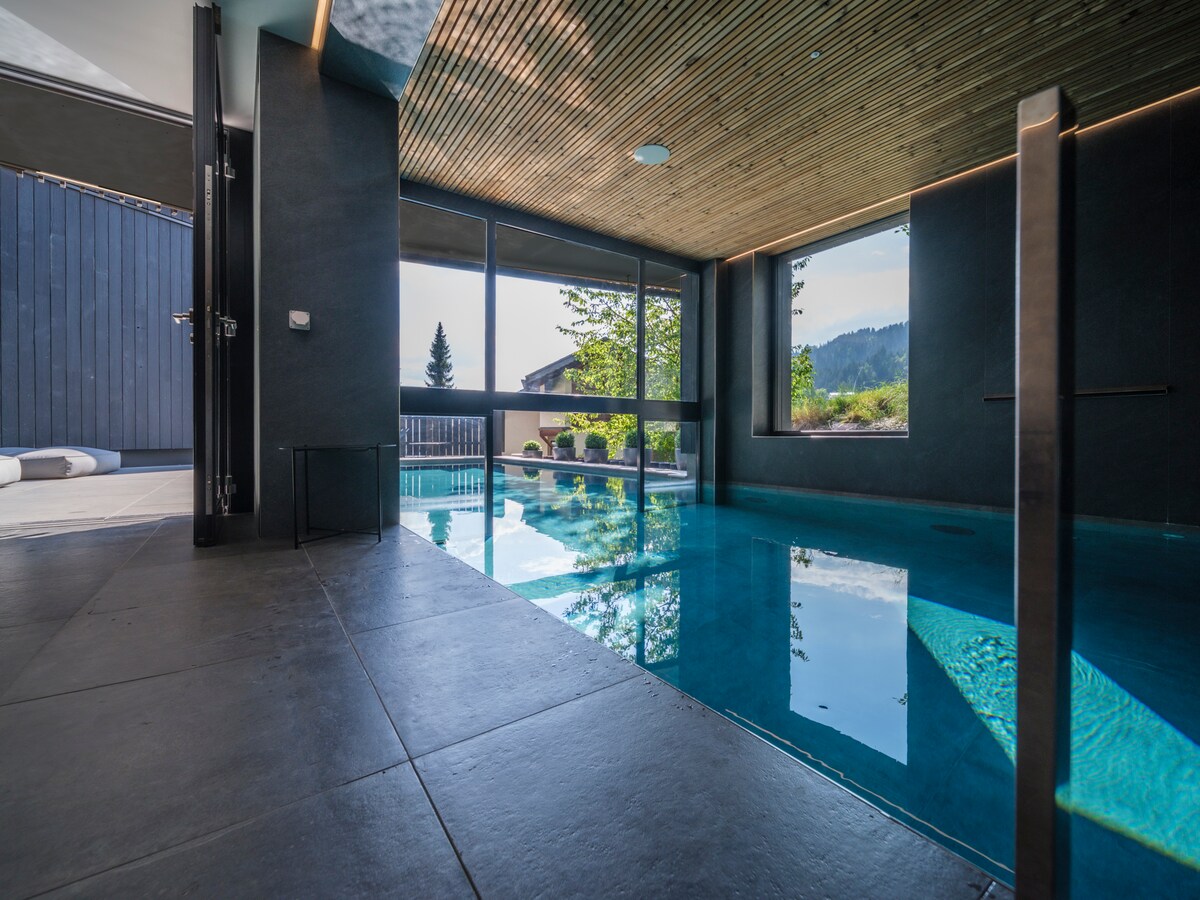 An indoor swimming pool features clear blue water, surrounded by large windows that offer views of the outdoors. The sleek design includes a wooden ceiling and tile flooring, creating a modern and tranquil atmosphere.