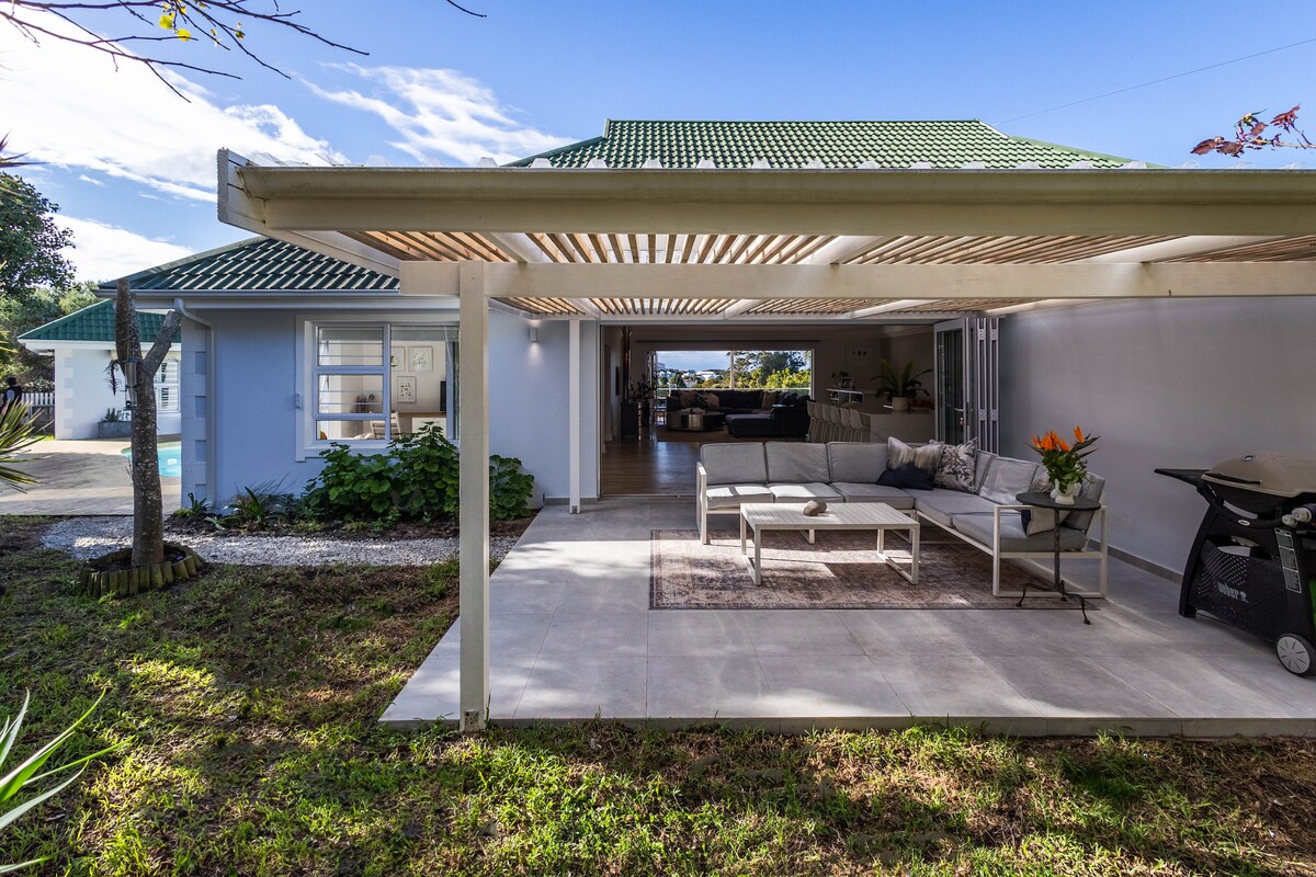 A spacious outdoor patio area features a stylish seating arrangement with light-colored cushions. The setting is complemented by a shaded pergola, and lush greenery surrounds the space. A glimpse of the pool area is visible in the background, enhancing the tranquil atmosphere.
