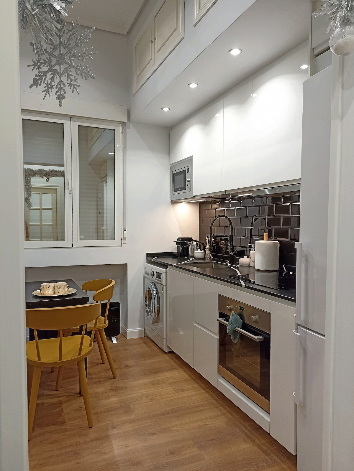 A modern kitchen area features sleek white cabinetry and stainless steel appliances. A small table with two yellow chairs is positioned beside a window, allowing natural light to brighten the space. Essential kitchen items, including a washing machine, are visible, highlighting the room's functionality.