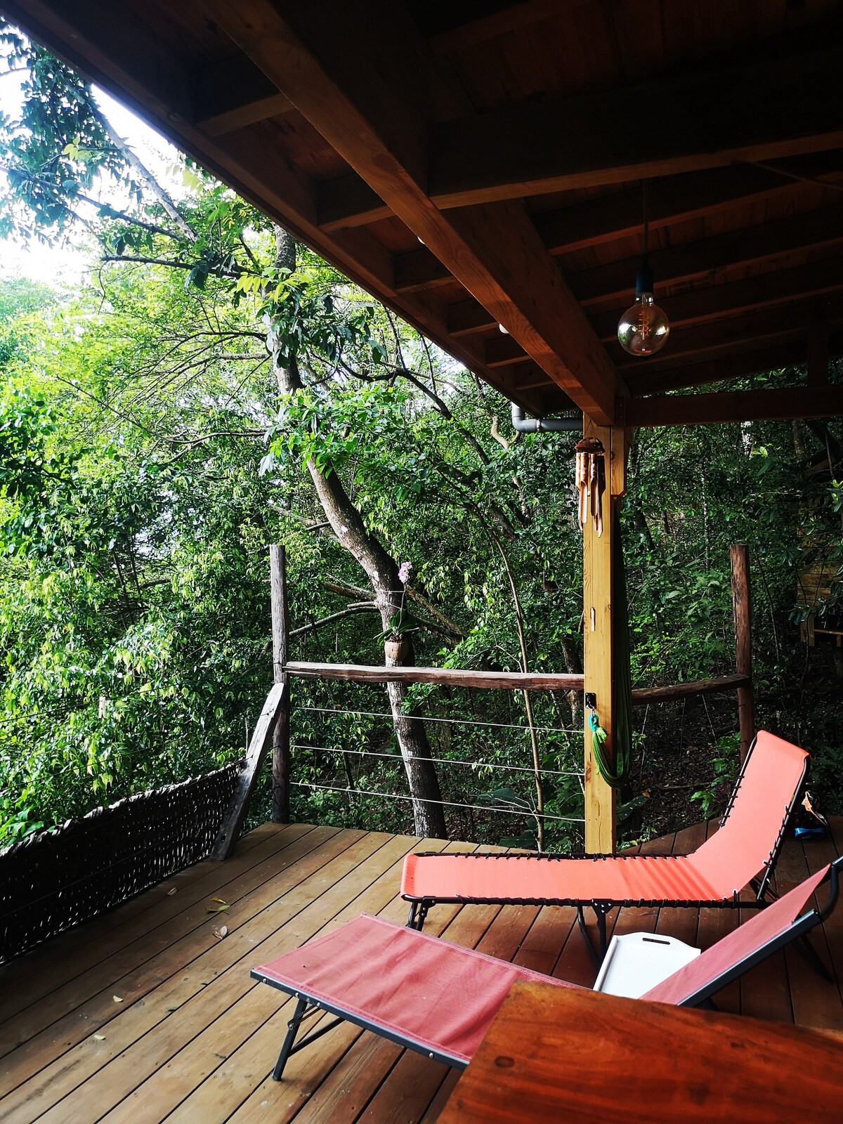 A wooden deck overlooks the lush greenery of the surrounding forest. Two lounge chairs are positioned for relaxation, inviting guests to enjoy the tranquility. The structure features wooden beams and a hanging light fixture, providing a cozy outdoor space immersed in nature.