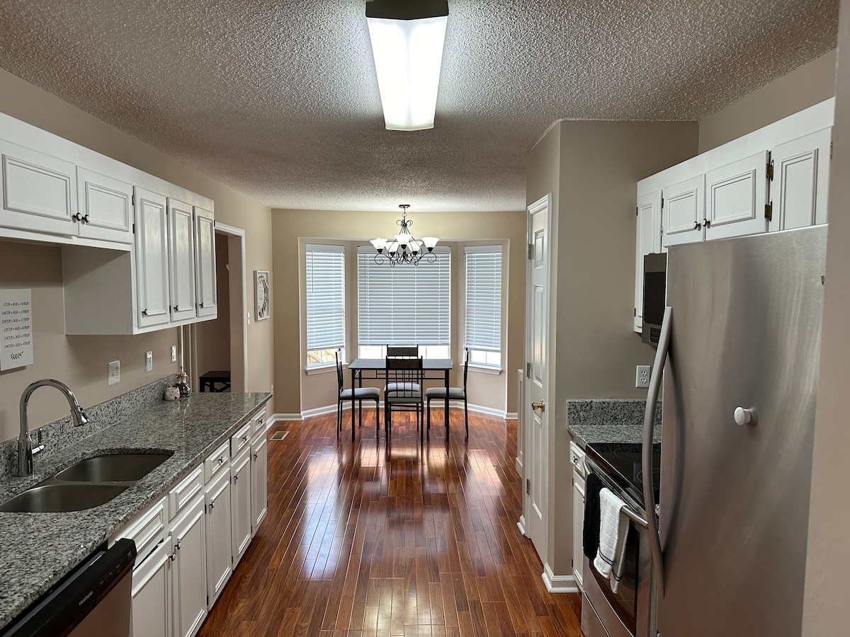 The kitchen features modern appliances and ample counter space, complemented by white cabinetry. A dining area with a contemporary chandelier and a table for four is visible through a large window, filling the space with natural light. Warm wood flooring adds a welcoming touch.