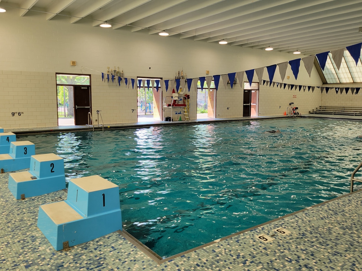 An indoor swimming pool is presented with clear blue water, surrounded by a tiled deck. Folding chairs are arranged nearby, with blue and white flags hanging overhead. Large windows allow natural light to fill the space, providing views of an outdoor area.