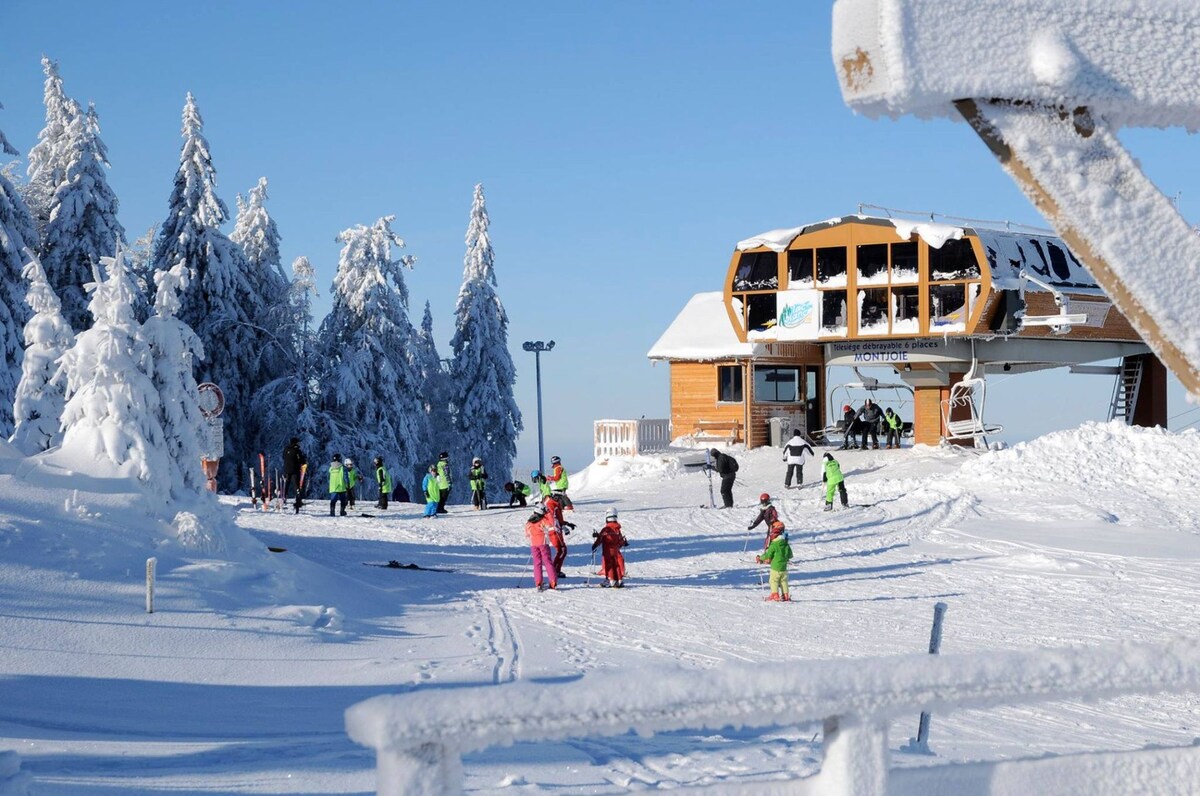 A winter scene at a ski resort features children and adults engaged in skiing and snowboarding on a snow-covered slope. A wooden ski lift station stands prominently in the background, surrounded by snow-draped trees, while a clear blue sky enhances the bright atmosphere.