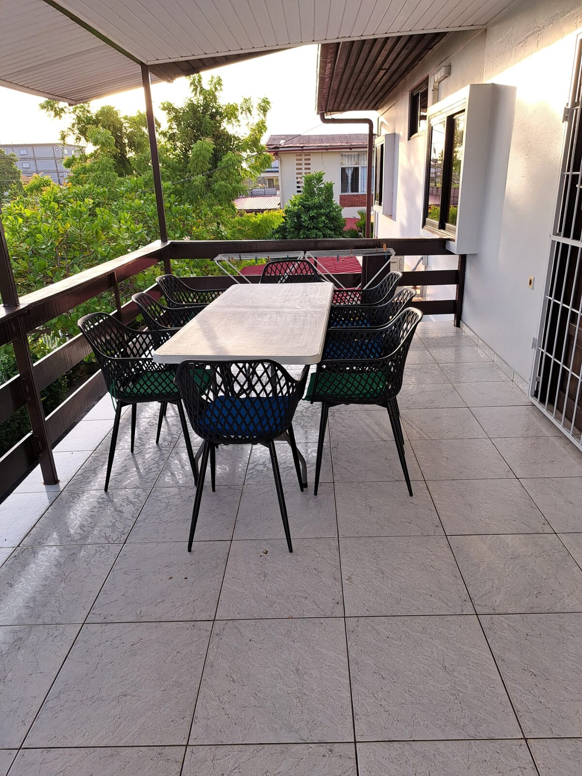A balcony area is presented with a large table surrounded by six black wicker chairs. The floor features light-colored tiles, and greenery can be seen through the railing, enhancing the outdoor space's inviting atmosphere.