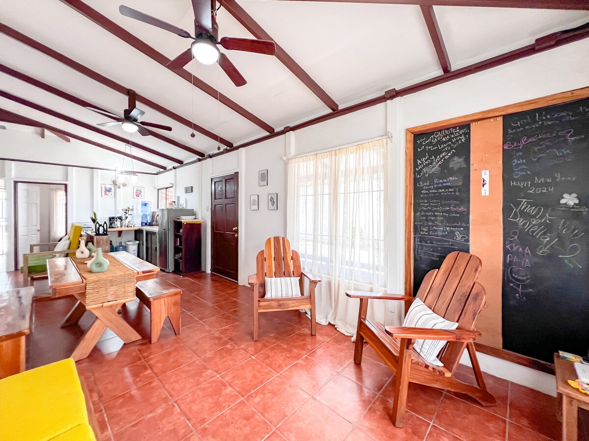 A spacious living area features wooden beamed ceilings and terracotta tile flooring. Two comfortable wooden chairs are positioned beside a large table. A chalkboard wall displays an array of colorful writings, while a window allows natural light to brighten the room.